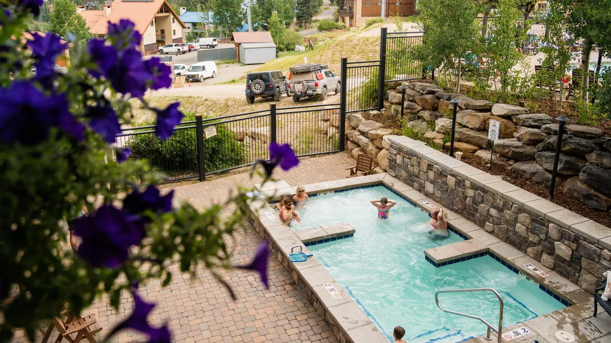 This image features a sunny outdoor pool surrounded by flowering plants, with people enjoying the water and lounging poolside. The overall mood is bright, social, and inviting, suggesting an ideal spot for relaxation and recreation during warmer months.