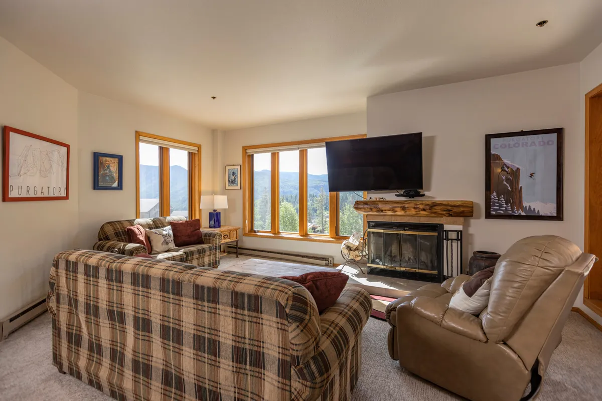 A warmly lit living room featuring a plaid sofa, a large window offering mountain views, a brick wood-burning fireplace, and a TV. Decorative art brings character to the space, while the arrangement encourages relaxing with family, watching movies, or cozying up by the fire.