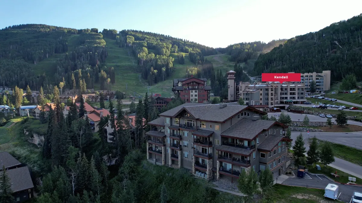 A panoramic view of mountain peaks rising above lush green trees, with charming residential buildings nestled at their base. The scene includes a parking area in the foreground and clear skies, suggesting calm and adventure. Activities like skiing, hiking, and mountain biking are possible here, while seating areas hint at relaxing moments outdoors.