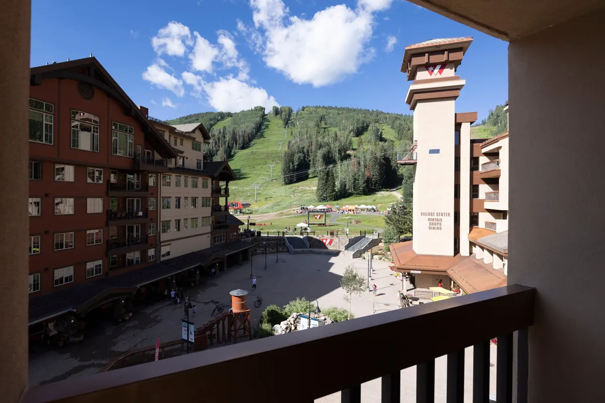 The image offers a vibrant view of a mountain ski lift, bustling outdoor plaza, and surrounding resort buildings, all set beneath expansive mountain views. The plaza is clearly a center for gathering and social activity, hinting at the lively, communal aspects of resort life.