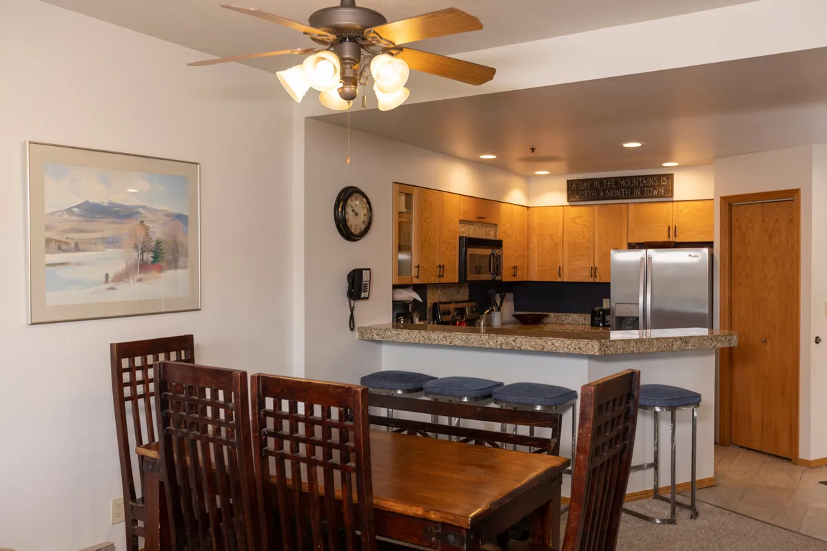 A bright, functional kitchen and dining space featuring a dining table, bar counter with stools, modern kitchen appliances, and tasteful wall art. The layout is ideal for preparing and sharing meals, with plenty of room for socializing.