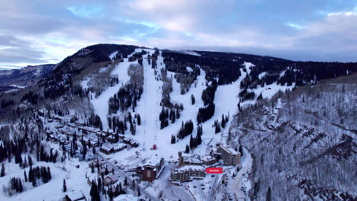 This image captures a sweeping view of snow-covered ski slopes framed by lodging buildings, ski lifts, and lush trees, all set against a dramatic mountain backdrop. The scene is lively, suggesting easy access from accommodation to the slopes and a range of winter activities.