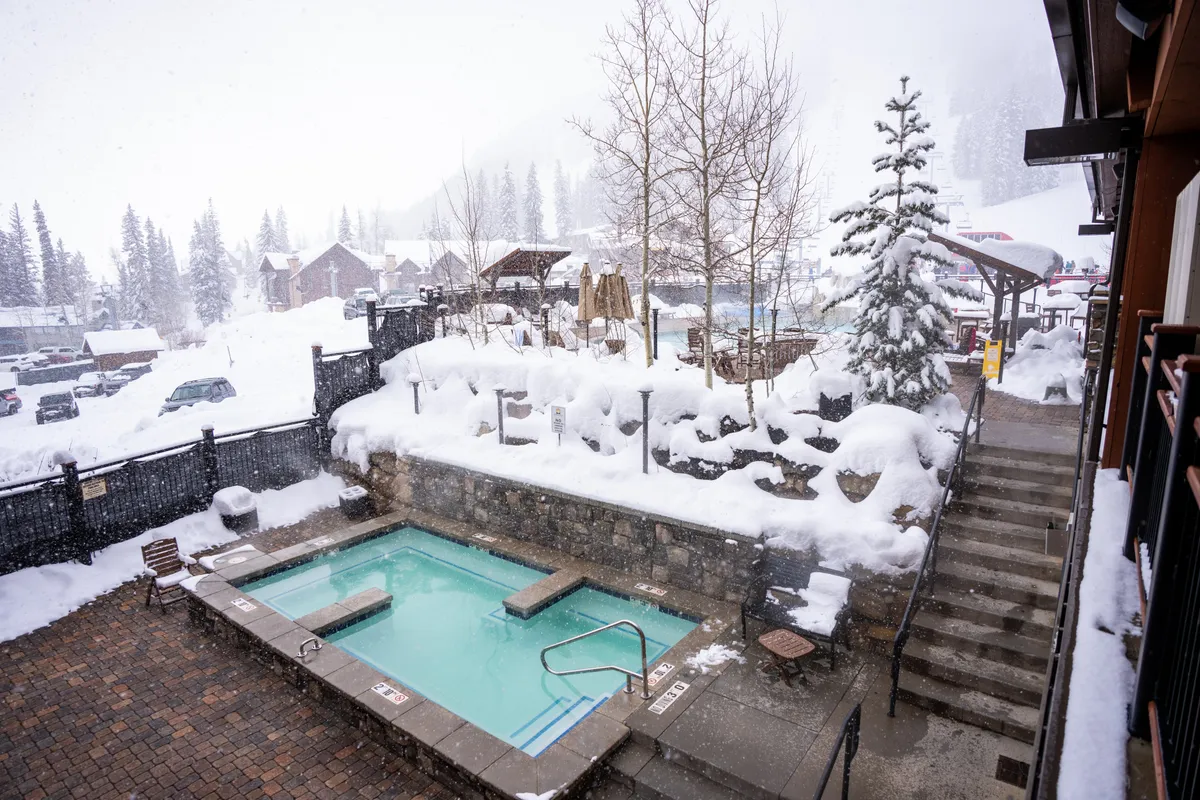 This image shows an outdoor hot tub surrounded by a snow-covered landscape, with mountains and trees providing a picturesque backdrop. The hot tub is steaming, suggesting a relaxing, warm experience even in winter. There are no people visible, keeping the focus on the amenity and its tranquil setting.