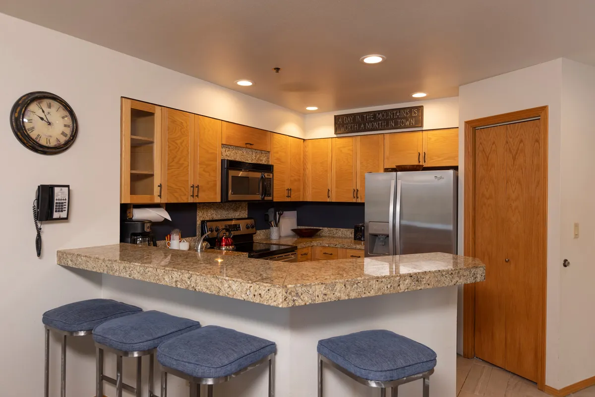 This image features a stylish kitchen with granite countertops, wooden cabinets, stainless steel appliances, bar stools, and a wall clock. The setup emphasizes both functionality and warmth, with space for cooking, entertaining, and casual dining at the breakfast bar.