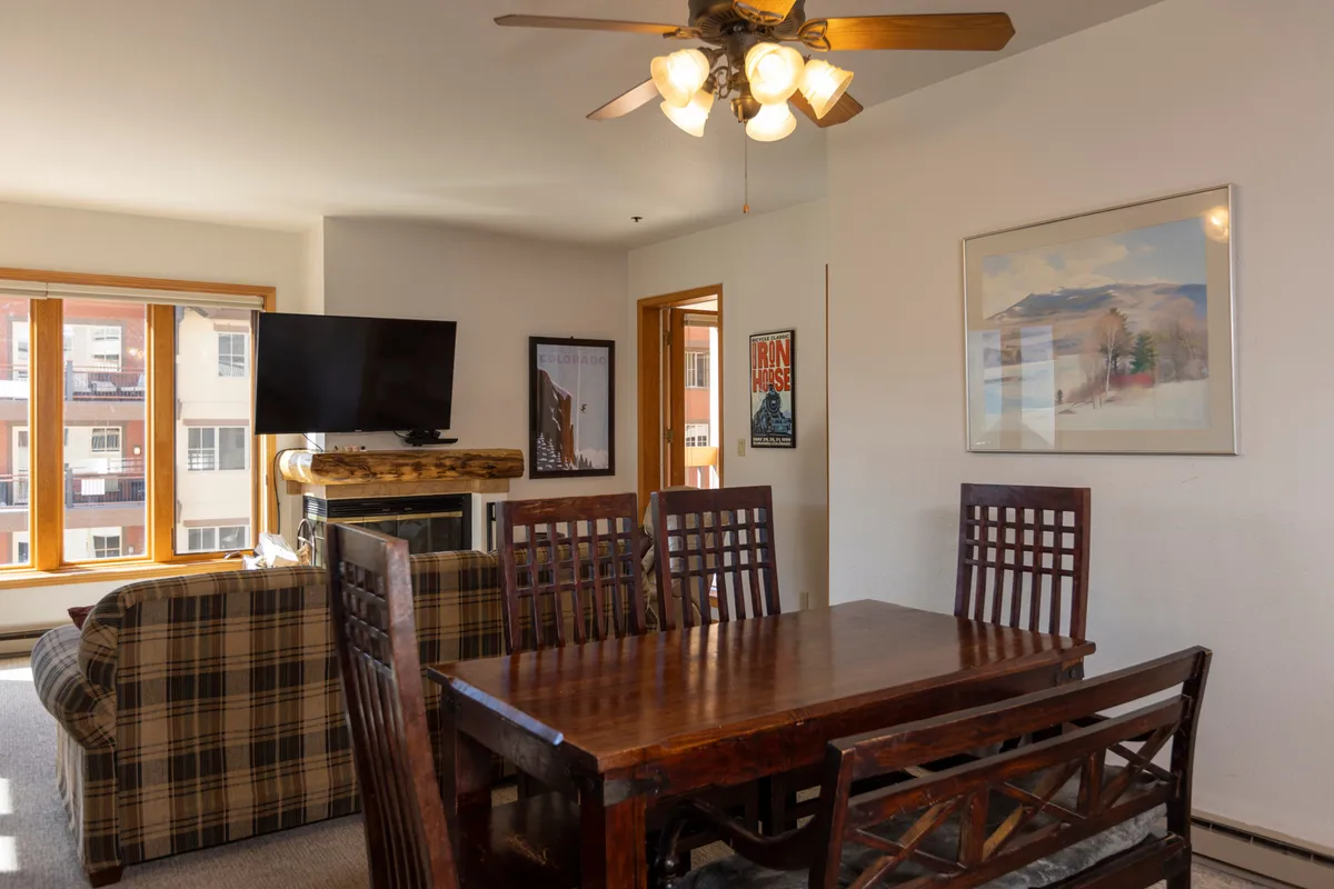 An inviting open-plan living and dining area, with a dining table set for meals and a plaid sofa near the fireplace. The television and framed artwork contribute to a welcoming, lived-in feel suitable for family activities, meals, and movie nights.