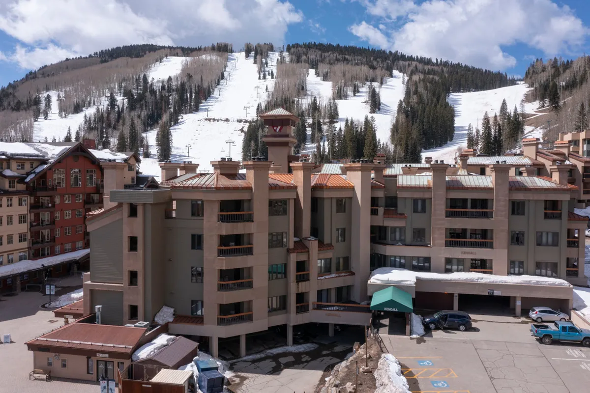 The exterior of a resort-style building surrounded by tall trees, with ski slopes in the background. Warm lighting and a gentle sunset glow create a cozy, inviting atmosphere, while hints of activity suggest the lively community and easy access to mountain adventures.