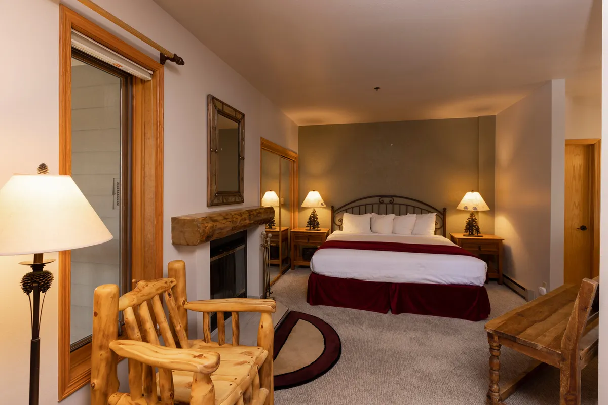 A cozy bedroom featuring a plush bed with layered blankets, a wooden chair beside it, soft ambient lamplight, a fireplace for warmth, and a decorative wall mirror. The gentle morning light streams in, creating a peaceful and inviting atmosphere ideal for rest.
